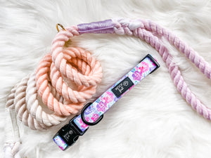Showcasing Flat lay image of  Pink and Purple flowers and green leaf pattern Dog Collar with metal D ring in black colour from Woof First. The Collar sitting on the tri-colour rope leash in colour pink, purple and grey. 